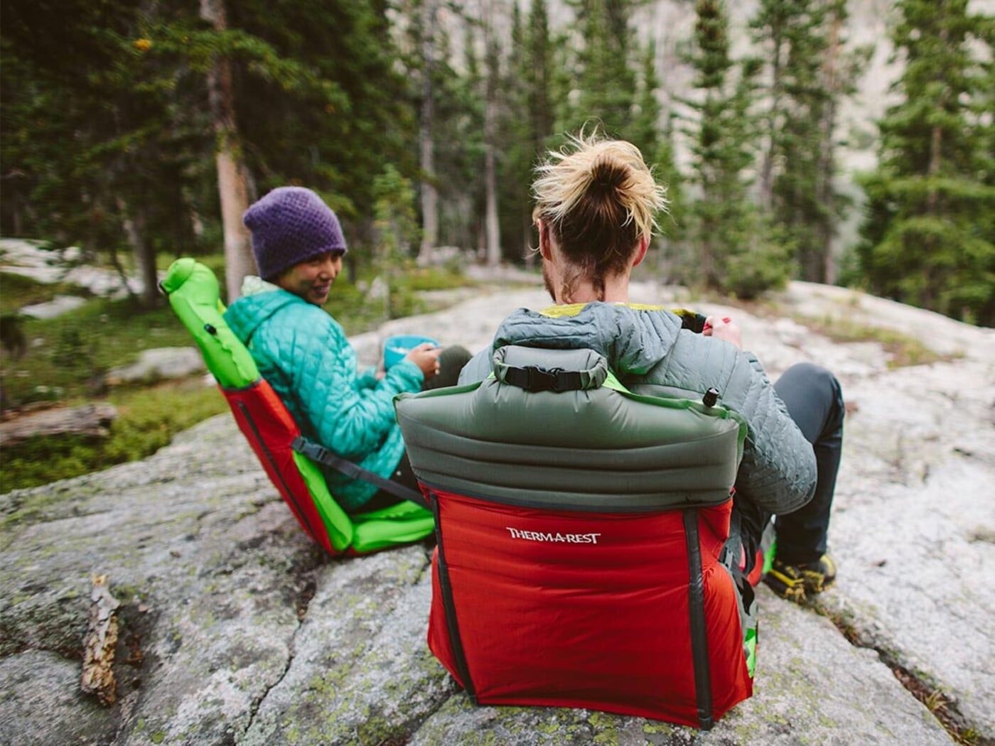 Two campers relaxing in pad-and-sleeve camp chairs on a rock above a forest
