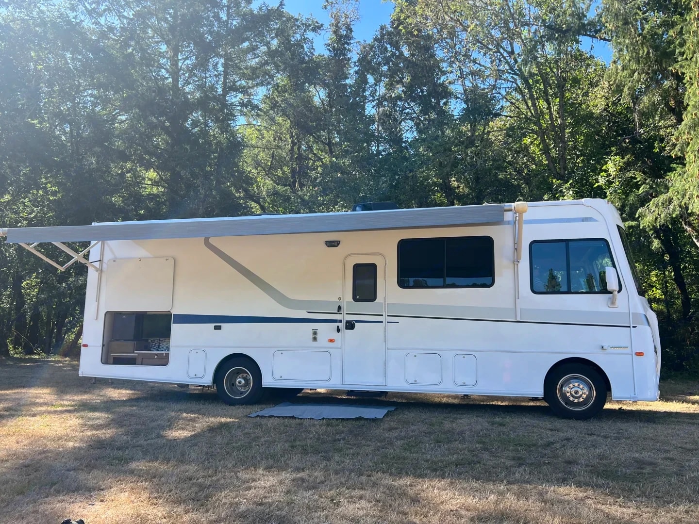 Passenger-side profile of the motorhome with awning out beside a treeline